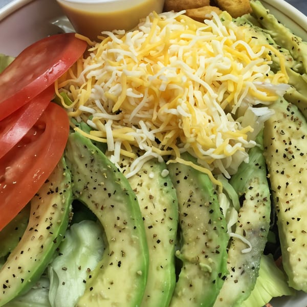 A Bull Chicks salad with sliced avocado, tomatoes, shredded cheese, and a side of dressing.